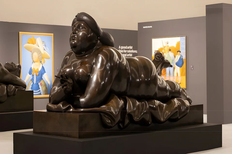 A bronze sculpture of a reclining woman on display in a gallery, with paintings visible in the background.