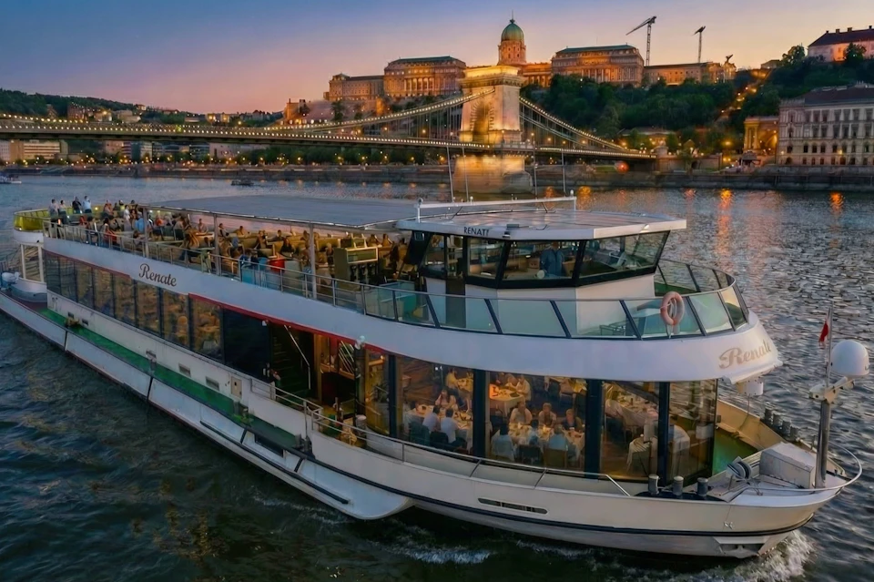 A riverboat with dining passengers cruises under the illuminated Chain Bridge at sunset, with a lit building in the background.
