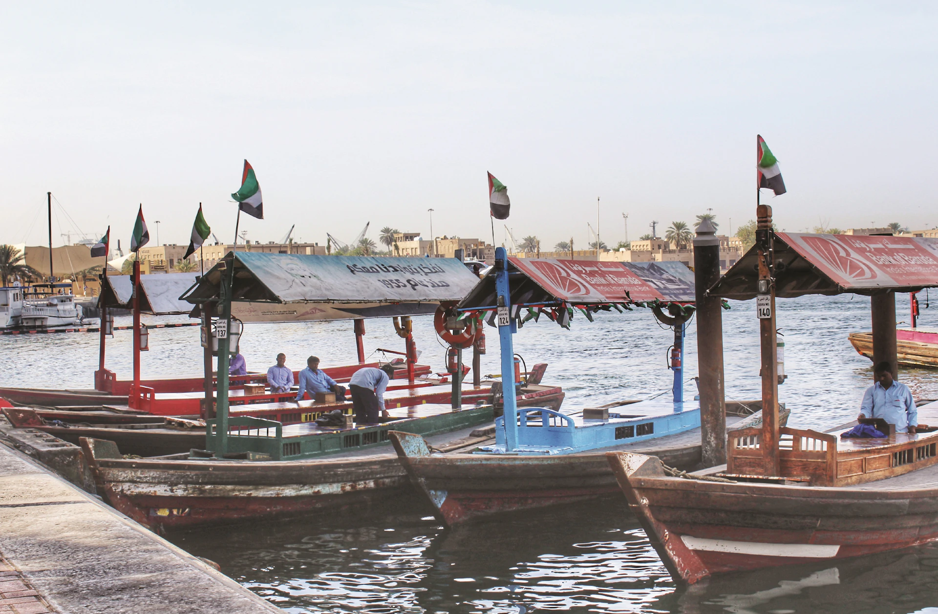 Small, colorful boats with awnings and flags docked at a waterside. Three people board a blue boat. Buildings and trees in the background.