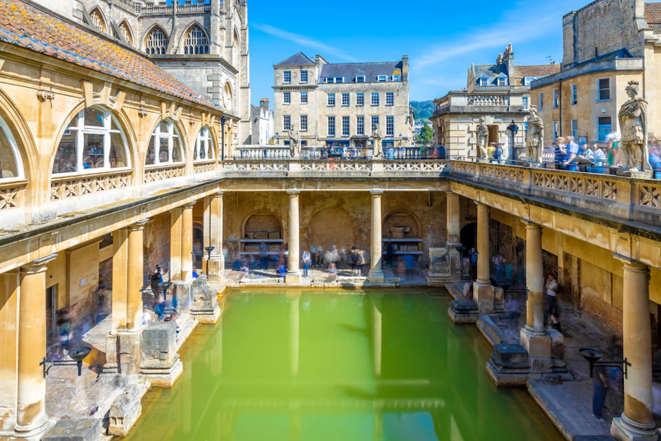 Historic Roman baths with green water surrounded by columns and arches, with people walking around; city buildings in the background.