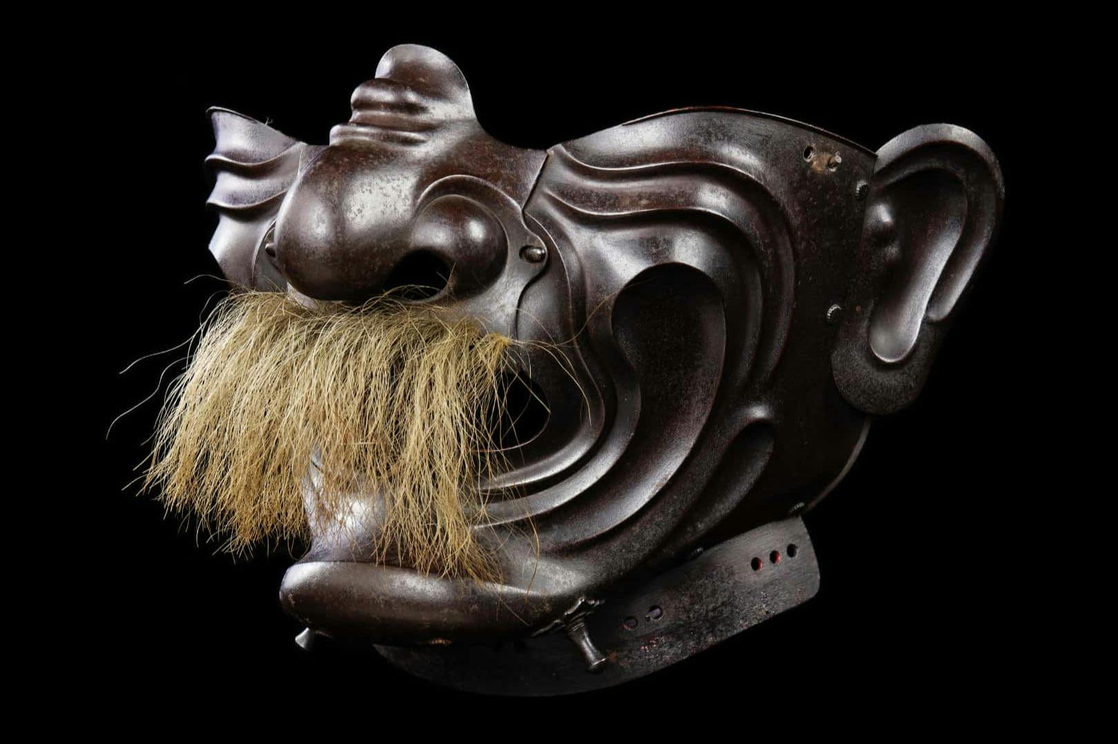 A traditional Japanese samurai mask with a large nose and a bushy mustache, made of metal, shown against a black background.