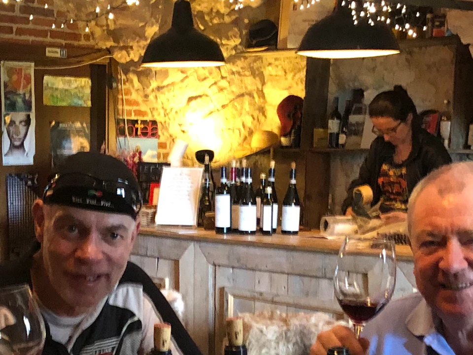 Four people sit around a wooden table with wine bottles, holding glasses in a cozy, rustic wine cellar adorned with string lights.