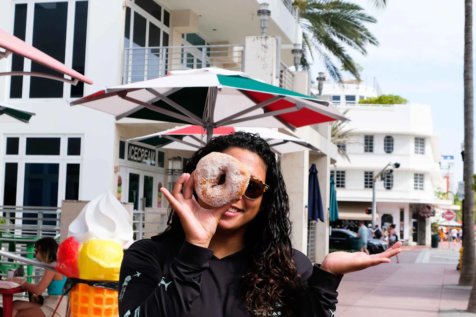 A woman holding a doughnut with a hole up to her eye, standing beside an ice cream cone statue outside an ice cream shop.