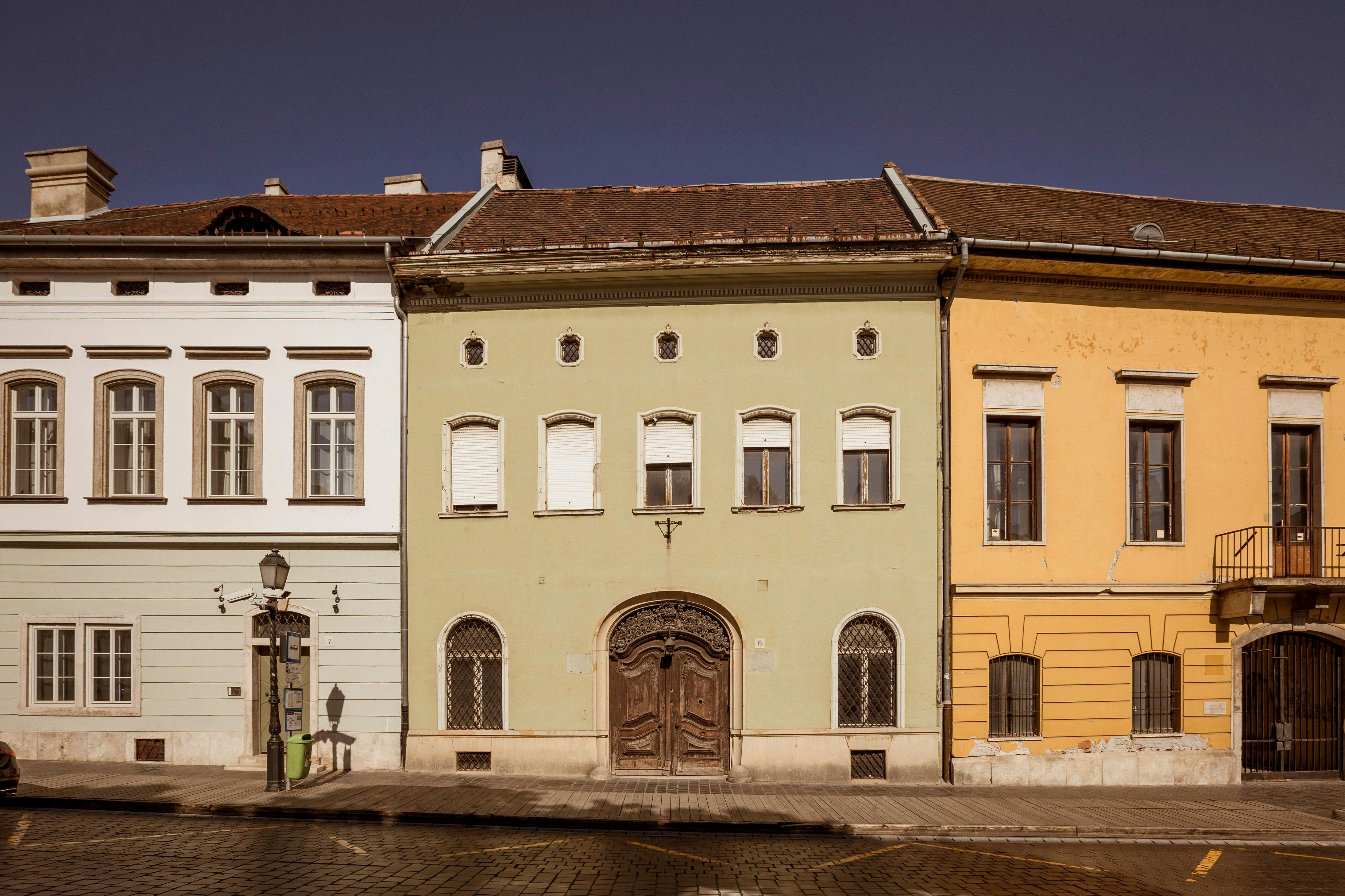 A row of old European buildings with distinct facades, one green with wooden doors, flanked by white and yellow ones.