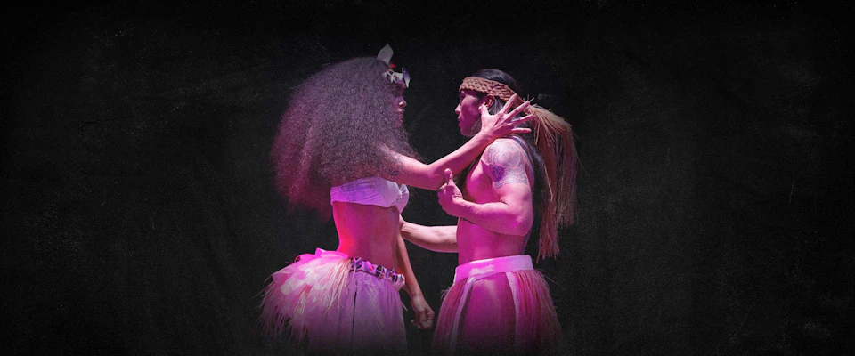 A man and woman in traditional attire perform a dance, illuminated by purple and pink lighting on a dark background.