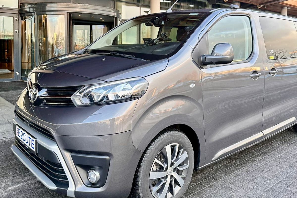 Gray minivan parked, showing front and side, with daytime running lights on. Entrance of a building is visible in the background.