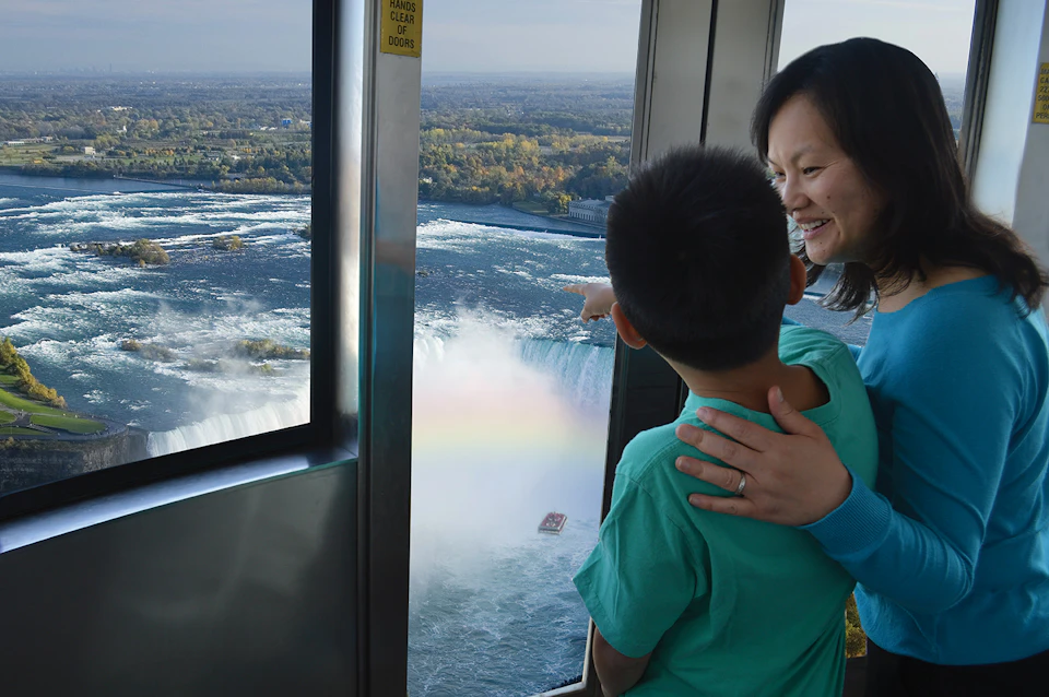 A woman and a child look out a window at Niagara Falls, with the child pointing at a rainbow over the waterfall.