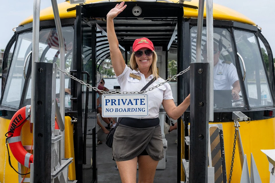 A woman in a uniform and red cap waves while standing at the entrance of a vehicle with a sign reading 'Private, No Boarding.'