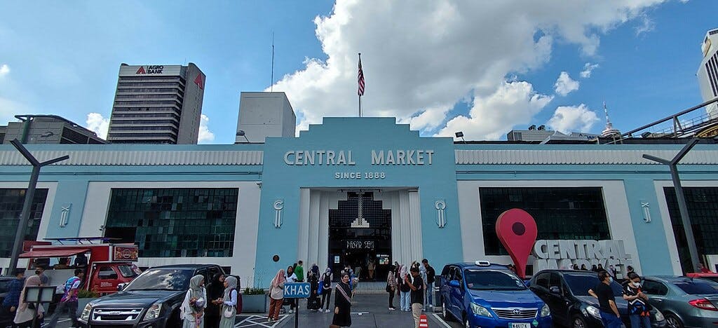 人们在中央市场（ Central Market ）前面，这是一栋蓝白相间的建筑，上面有一个大标志和停放的汽车。 天空部分多云。