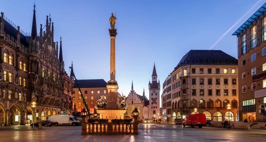 A city square at dawn with historic buildings, a tall column with a statue, and vehicles parked along the sides.