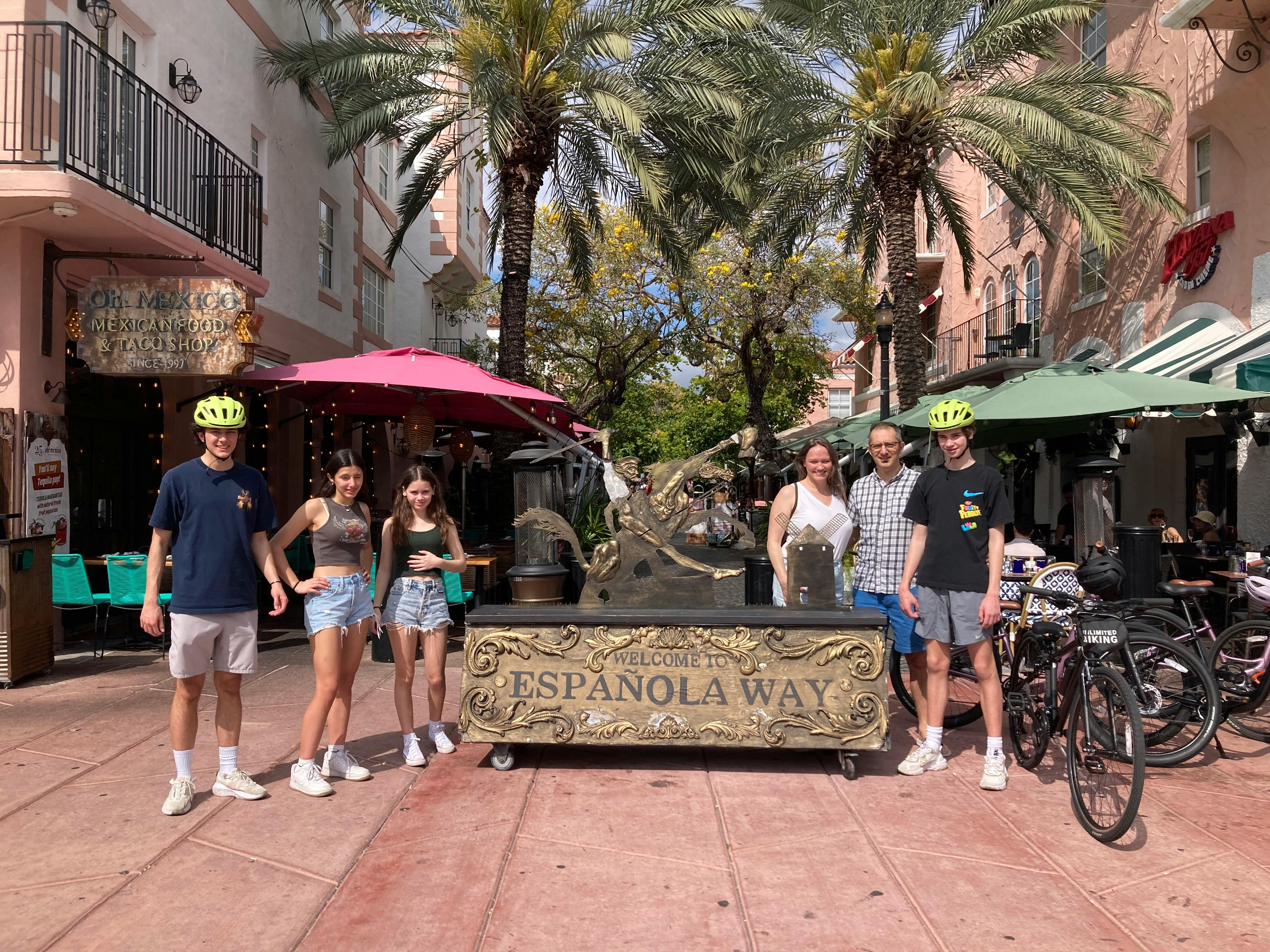 Skupina lidí se usmívá před nápisem „Welcome to Espanola Way“ s obchody, palmami a jízdními koly v pozadí.