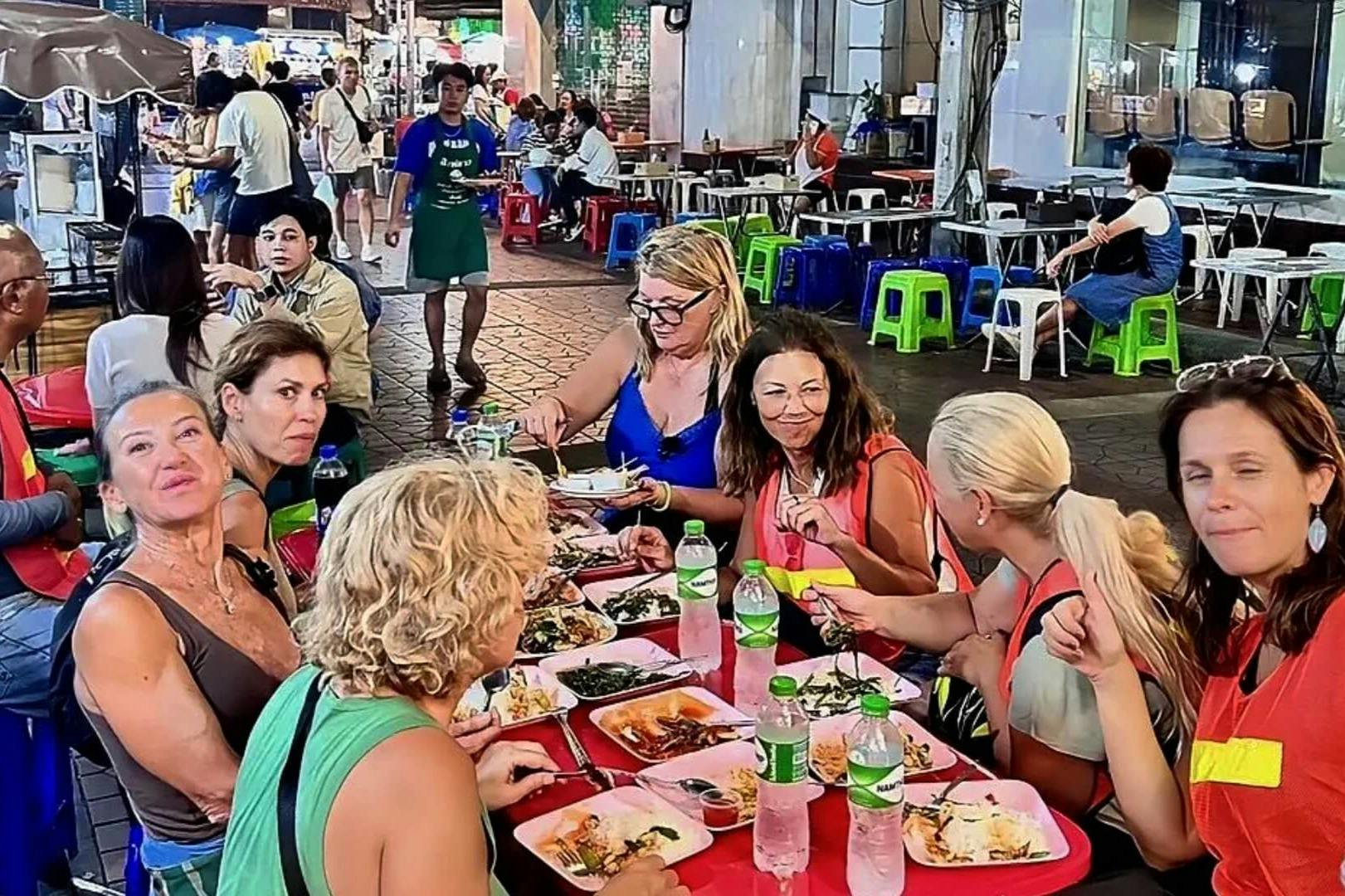 カラフルなテーブルと椅子が置かれた屋外の食料品市場で食事と飲み物を楽しむ女性のグループ。