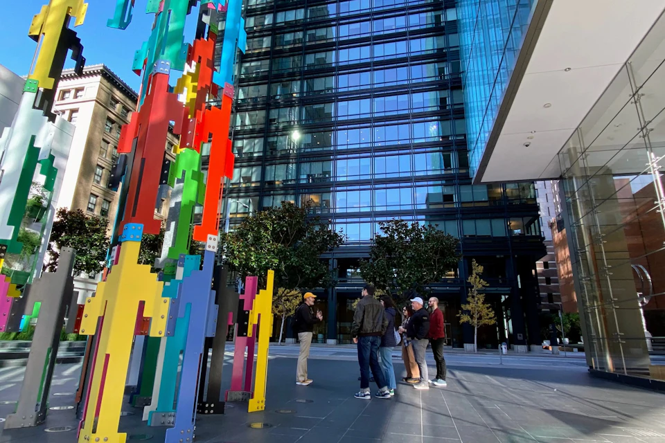 People stand near a colorful abstract sculpture in a city plaza, with a modern glass building in the background.