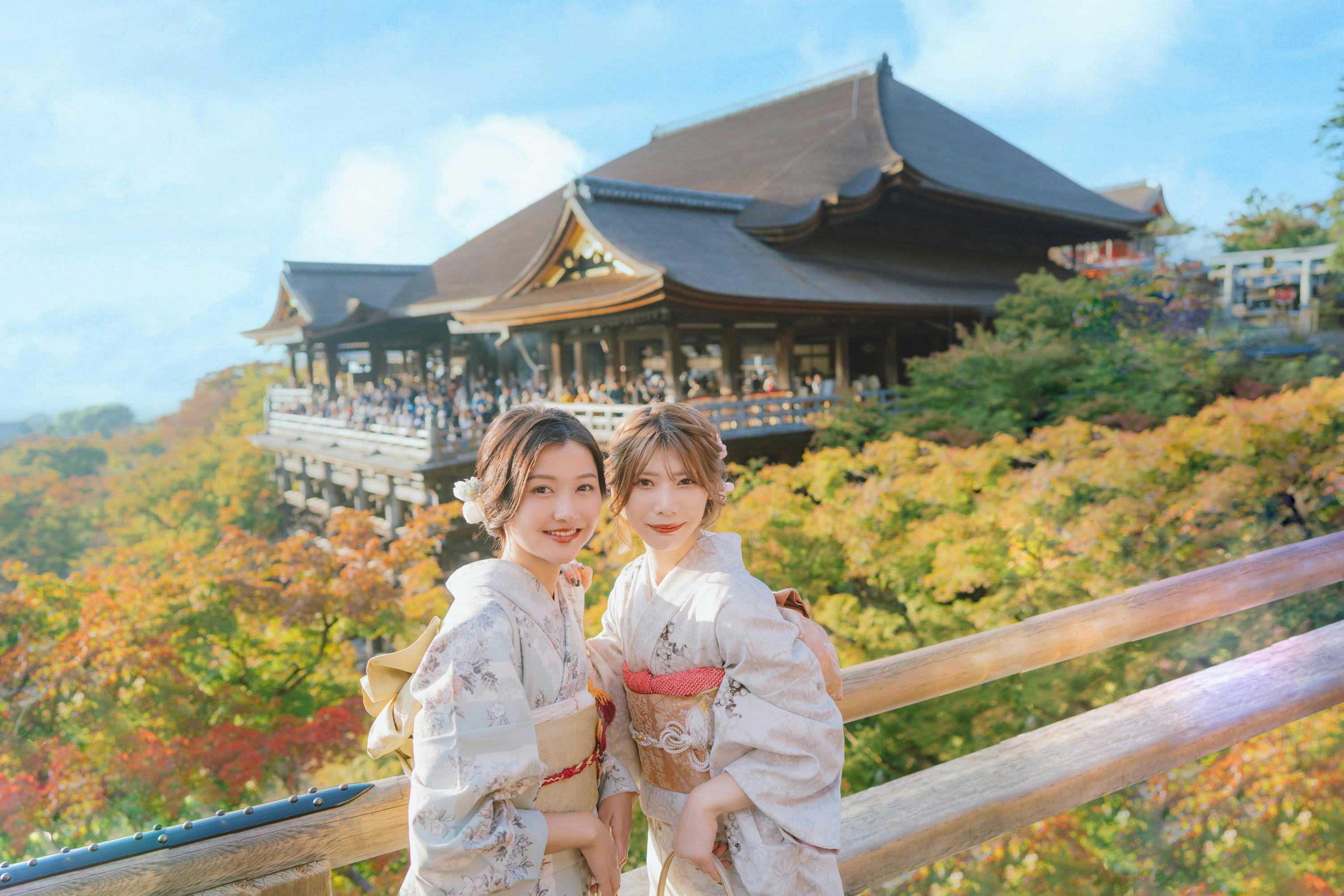 Two women in traditional kimonos posing in front of a historic wooden temple surrounded by colorful autumn foliage.