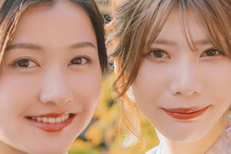 Two women in traditional kimonos posing in front of a historic wooden temple surrounded by colorful autumn foliage.