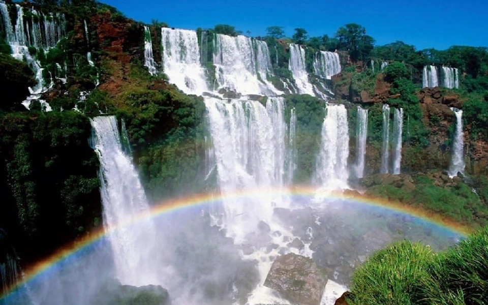 Multiple cascading waterfalls with lush greenery, under a clear blue sky. A rainbow arches through the misty spray.