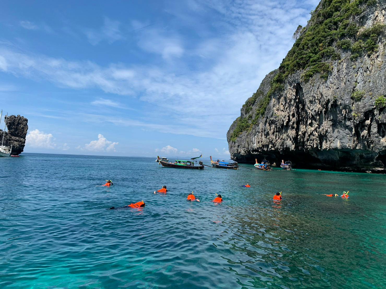 Des personnes portant des gilets de sauvetage orange font de la plongée avec tuba près de bateaux et de falaises rocheuses dans une eau bleue claire par une journée ensoleillée.