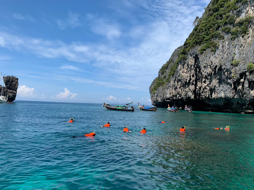 People in orange life jackets snorkeling near boats and rocky cliffs in clear blue water on a sunny day.