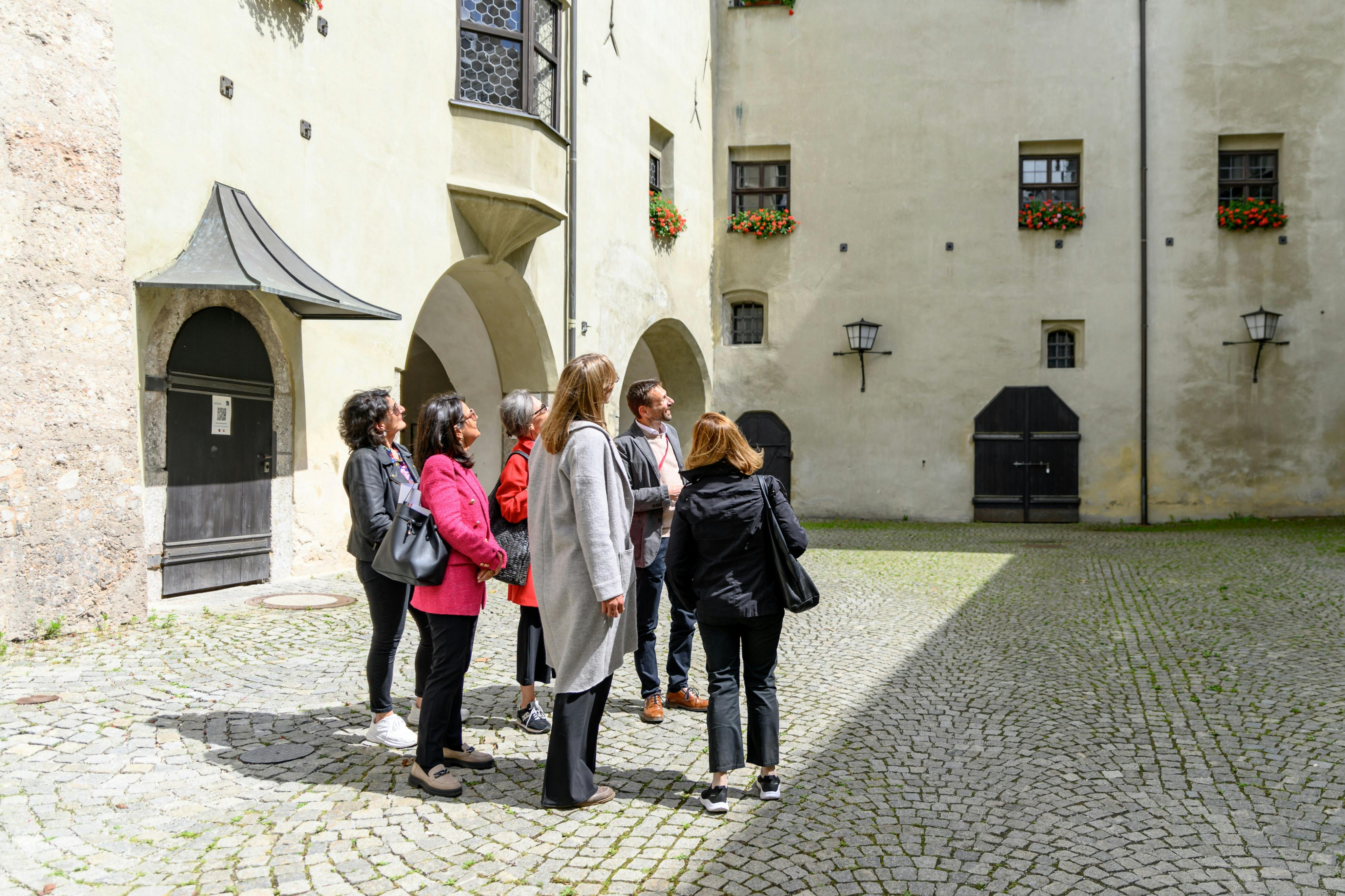 Группа людей стоит в булыжном дворе, окруженном историческими зданиями с арочными дверными проемами и цветочными коробками.