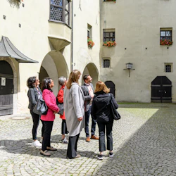 A group of people standing in a cobblestone courtyard surrounded by historic buildings with arched doorways and flower boxes.