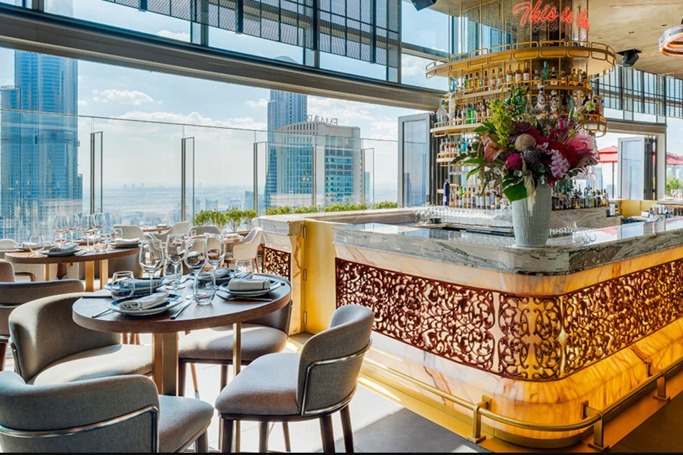 Elegant rooftop restaurant with a bar, dining tables, chairs, floral arrangement, and cityscape view through glass panels.