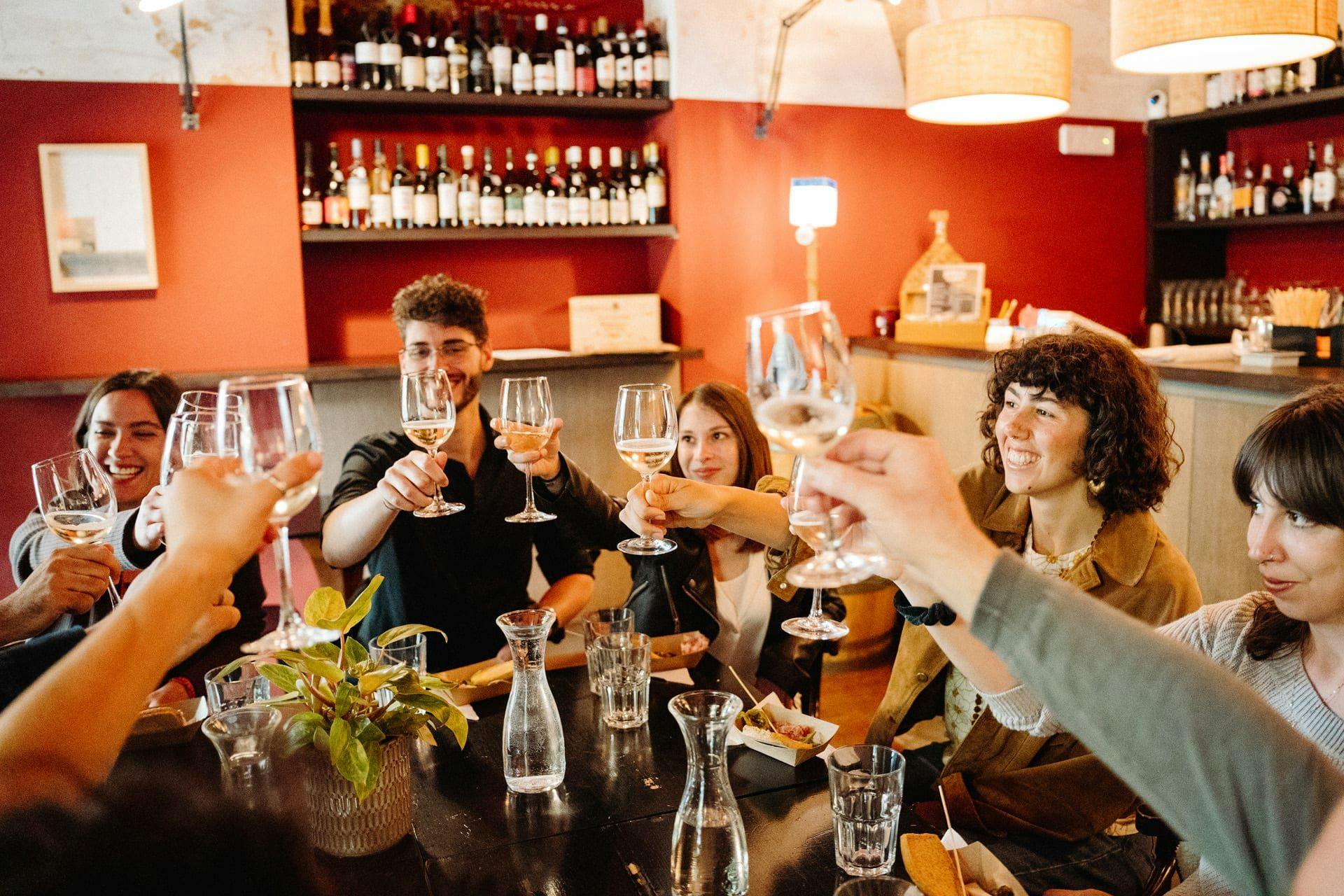 Group of people sitting at a restaurant table, raising wine glasses in a toast, with shelves of wine bottles in the background.