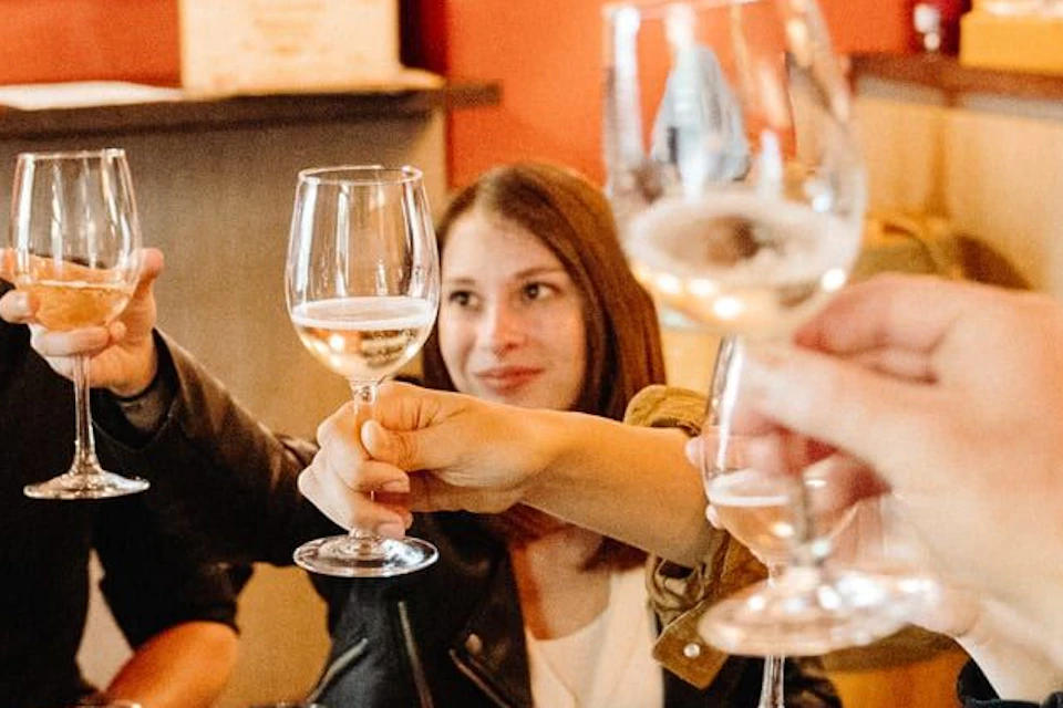 Group of people sitting at a restaurant table, raising wine glasses in a toast, with shelves of wine bottles in the background.