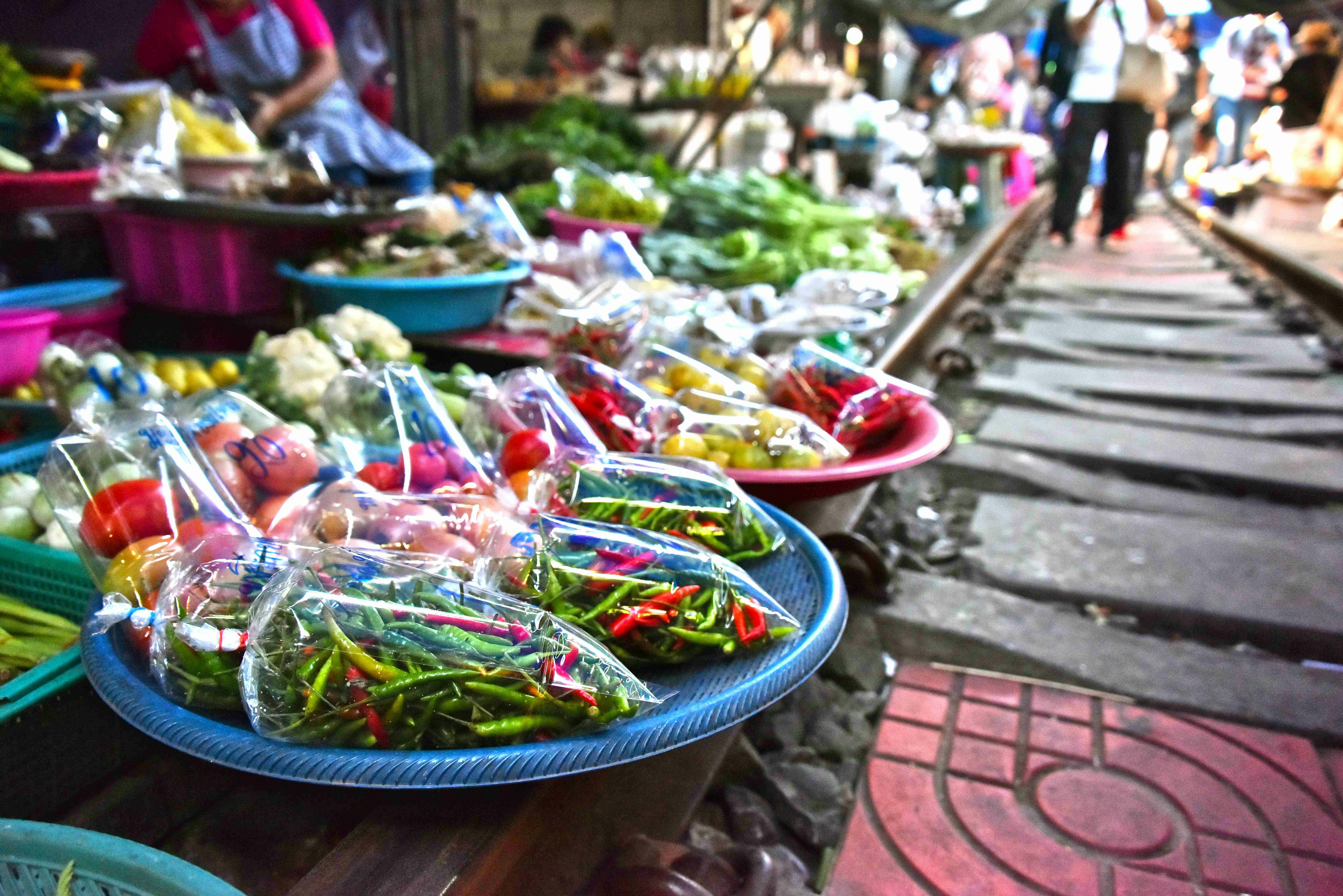 Fresh vegetables and herbs in plastic bags displayed on trays at a market stall next to train tracks.