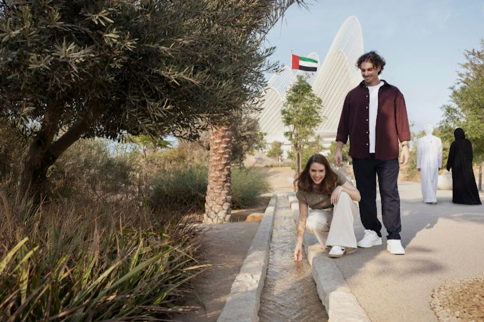 한 여성이 수로에서 물을 만지기 위해 웅크리고 있으며, 한 남성이 근처에 서 있습니다. 배경에는 현대적인 건물과 아랍에미리트 국기가 있습니다.