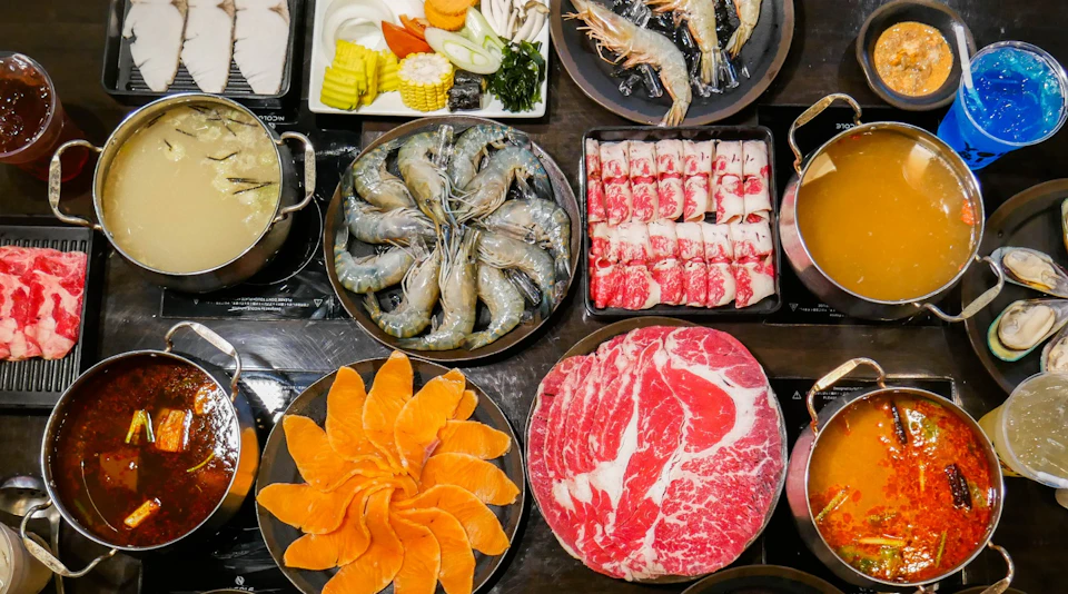 A table set with raw ingredients for hot pot, including shrimp, sliced meat, salmon, and vegetables.