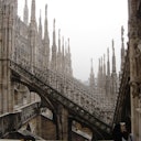 Gothic architecture with numerous ornate spires and flying buttresses on a cloudy day. People are visible at the bottom right.