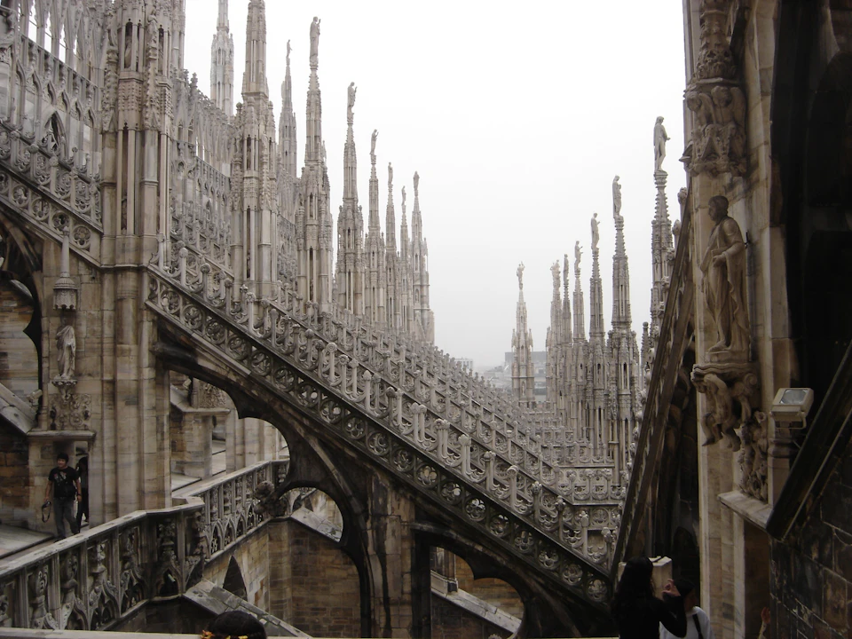 Gothic architecture with numerous ornate spires and flying buttresses on a cloudy day. People are visible at the bottom right.