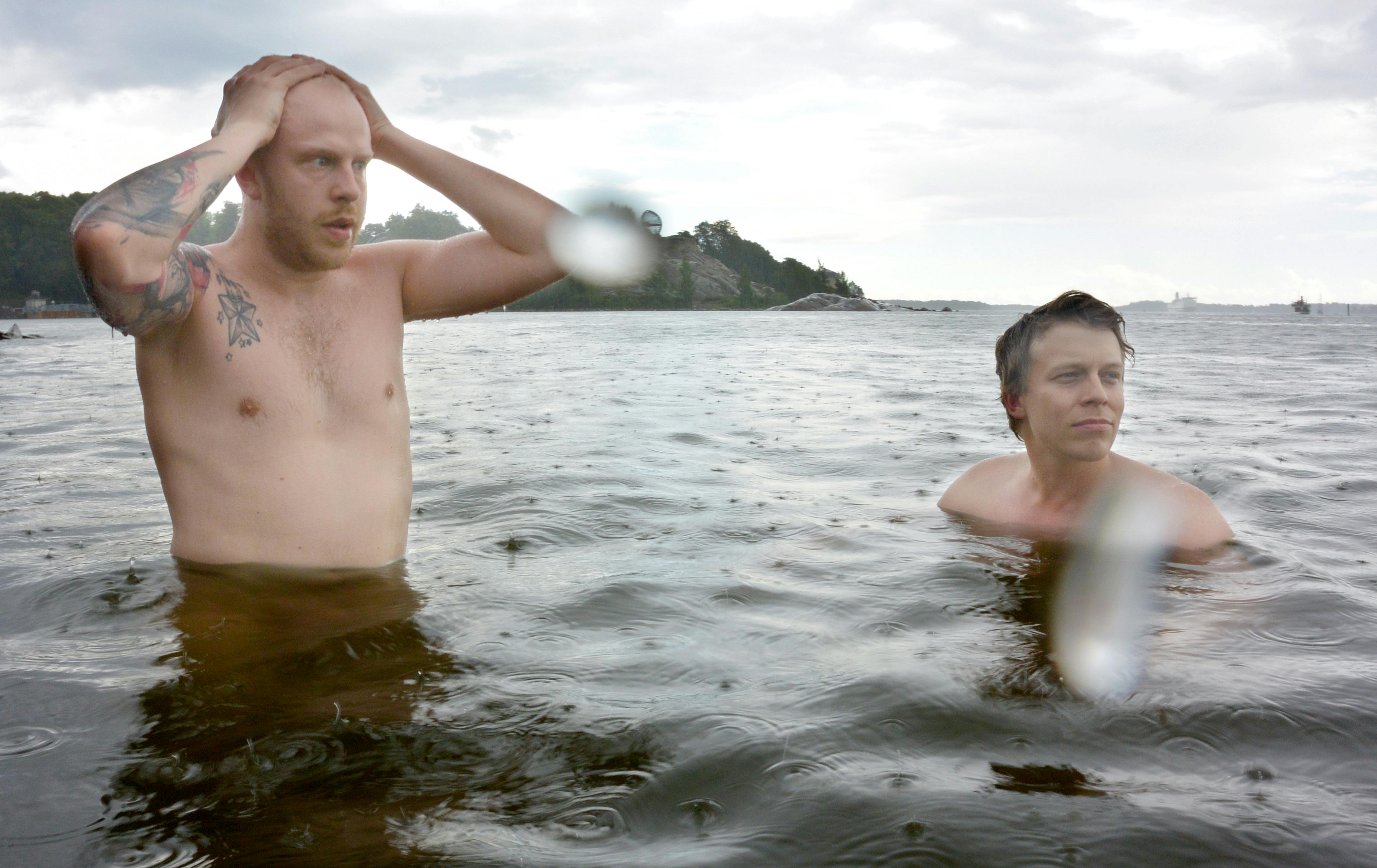 Δύο άνθρωποι χωρίς πουκάμισο στέκονται σε ρηχά νερά, με τον έναν να κρατά το κεφάλι τους. Στο βάθος φαίνεται ένα νησί.