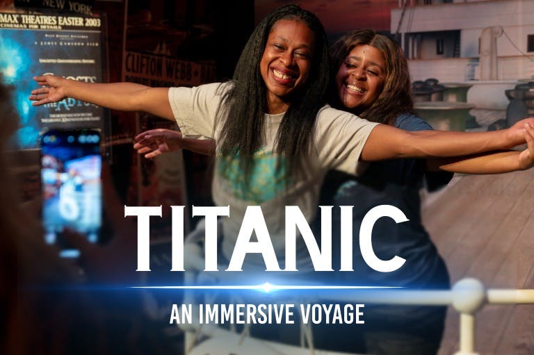 Två personer ler medan de poserar i en miljö med Titanic-tema, med en skylt där det står "Titanic: An Immersive Voyage."