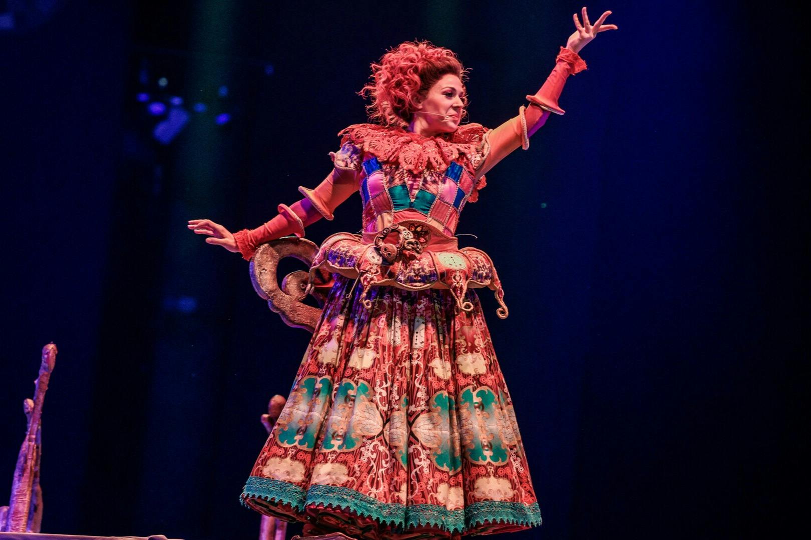 A performer in a colorful, elaborate costume poses dramatically on stage against a dark background.