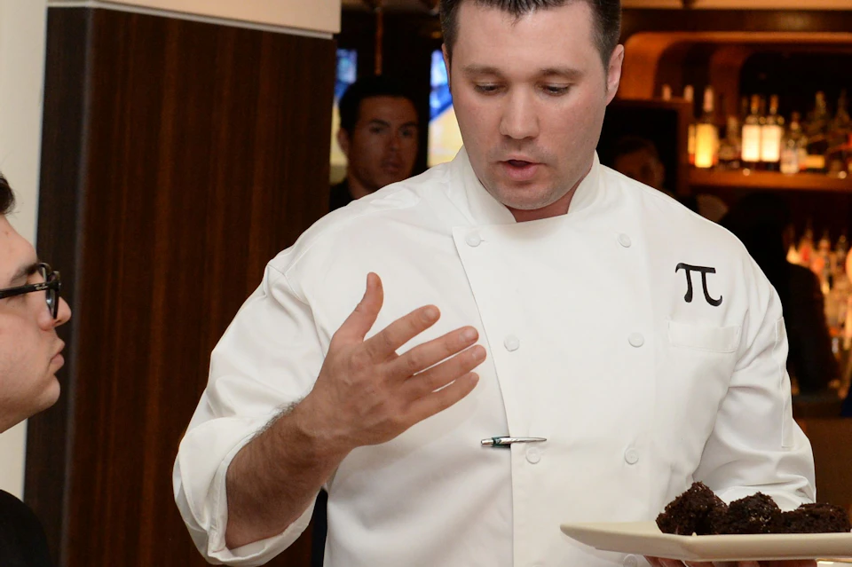 A chef in a white coat holding a plate of desserts, interacting with seated guests in a restaurant setting.
