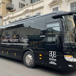 Black tour bus for Evan Evans Tours, parked on a street in front of beige buildings, advertising London to Bicester Village route.