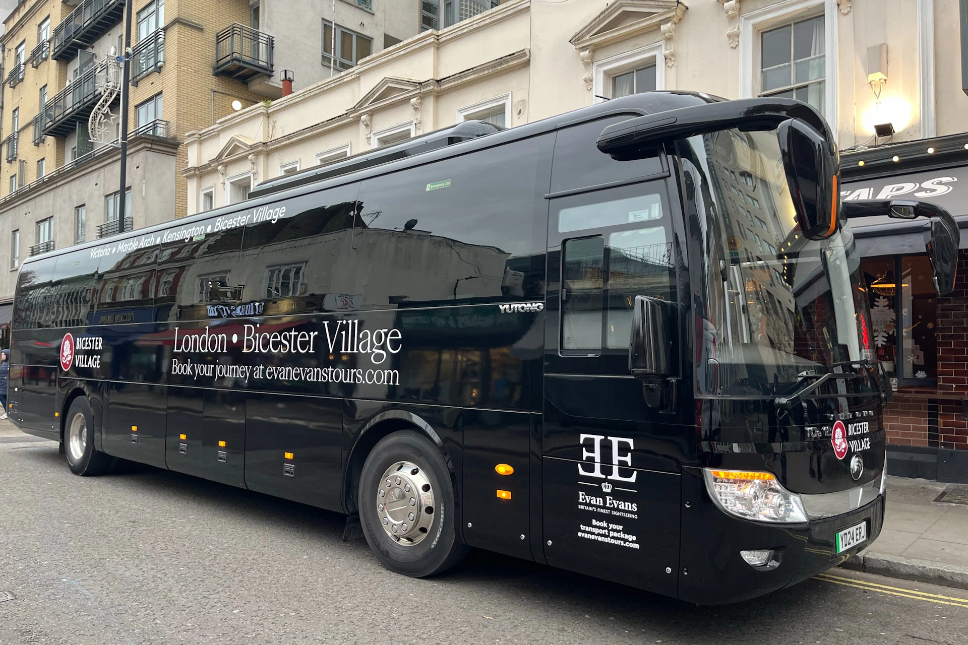 Black tour bus for Evan Evans Tours, parked on a street in front of beige buildings, advertising London to Bicester Village route.