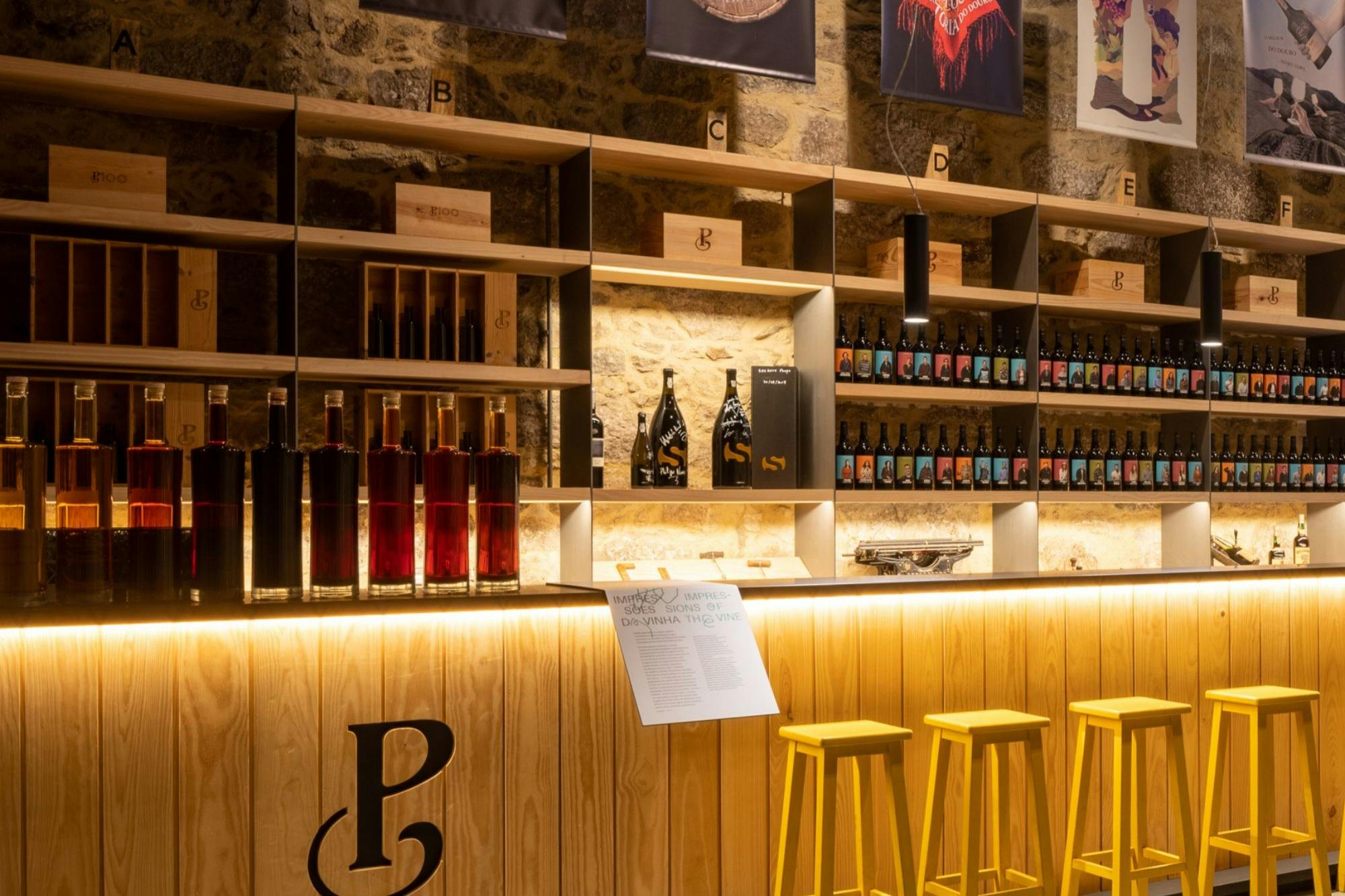 Een stijlvolle bar met houten planken met wijnflessen, een verlichte toonbank met gele krukken en een warm interieur.