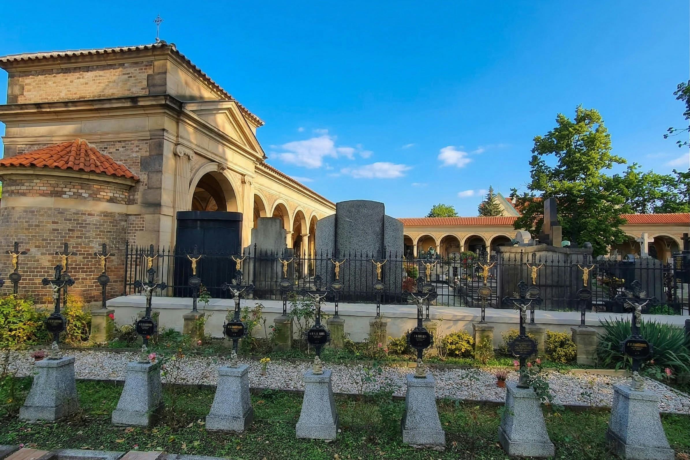 A cemetery with headstones, a wrought iron fence, and an ornate building with arches under a clear blue sky.