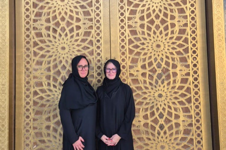 Δύο άνθρωποι που φορούν μαύρα ρούχα και μαντίλες στέκονται μπροστά από μια περίτεχνη, χρυσή πόρτα με περίπλοκα γεωμετρικά μοτίβα.