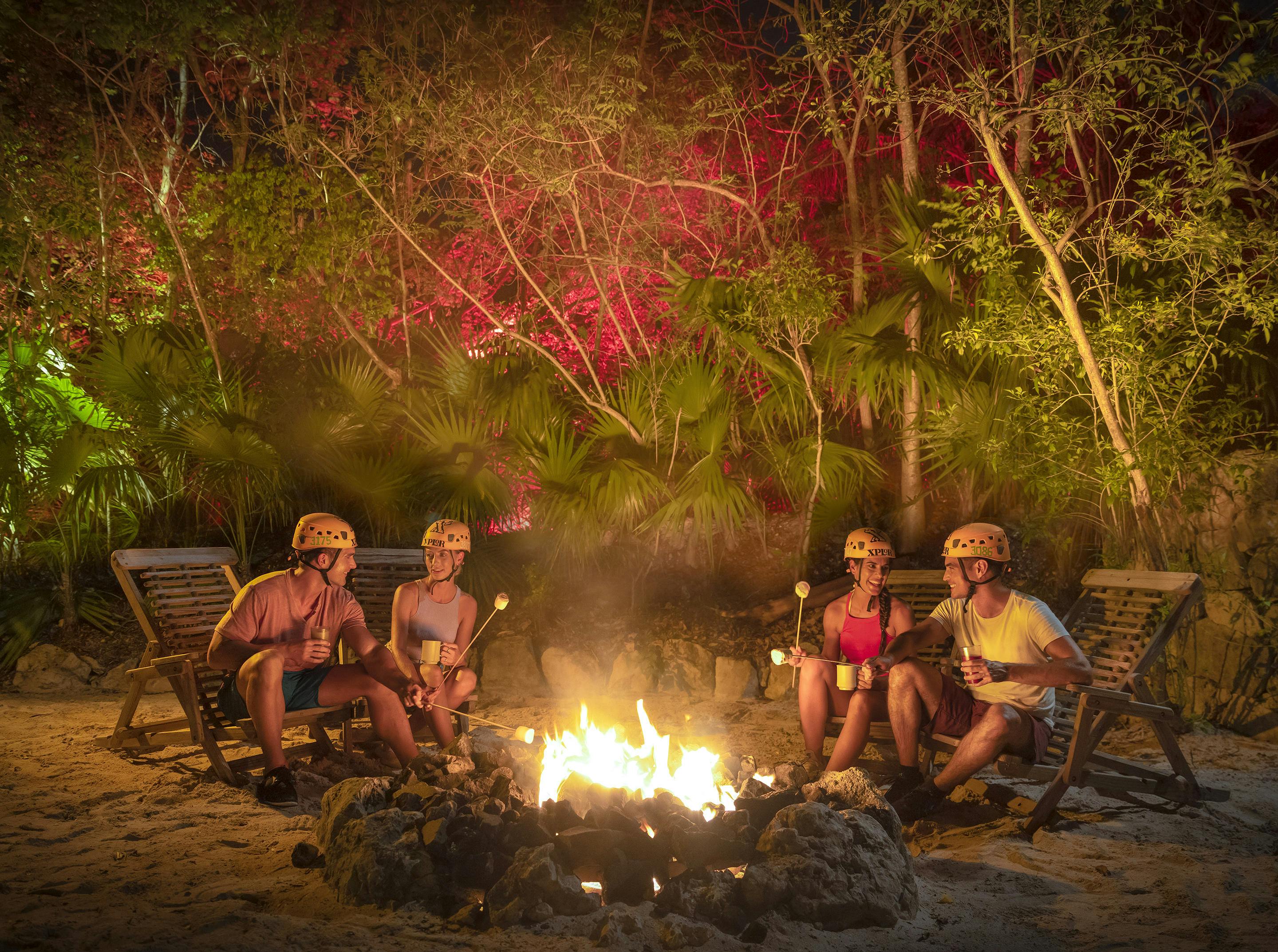 Cuatro personas con cascos asando malvaviscos alrededor de una hoguera por la noche, rodeadas de árboles con iluminación de colores.