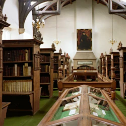 Ett historiskt bibliotek med bokhyllor i trä, glasfodral, ljuskronor och ett porträtt som hänger på bakväggen.