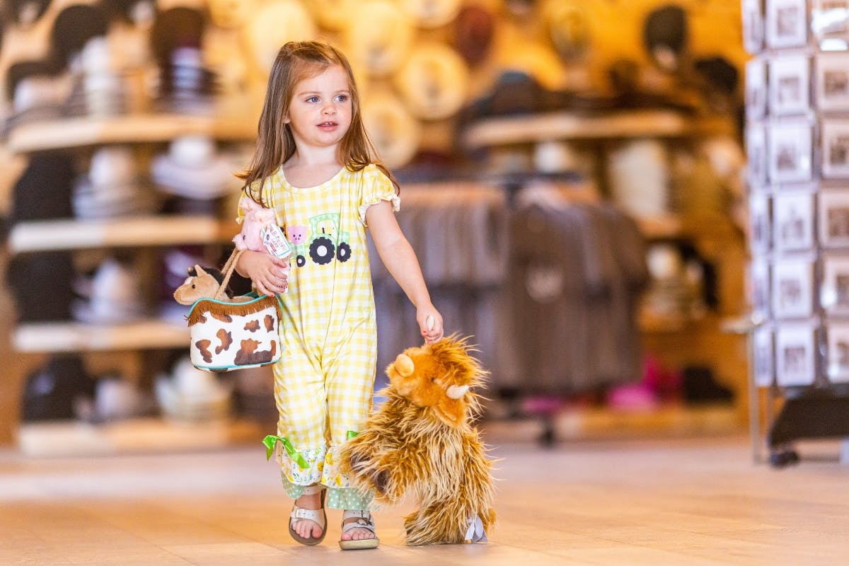 A young girl in a yellow checkered outfit holds a plush toy and a small cow-patterned handbag, standing in a shop with hats on shelves.