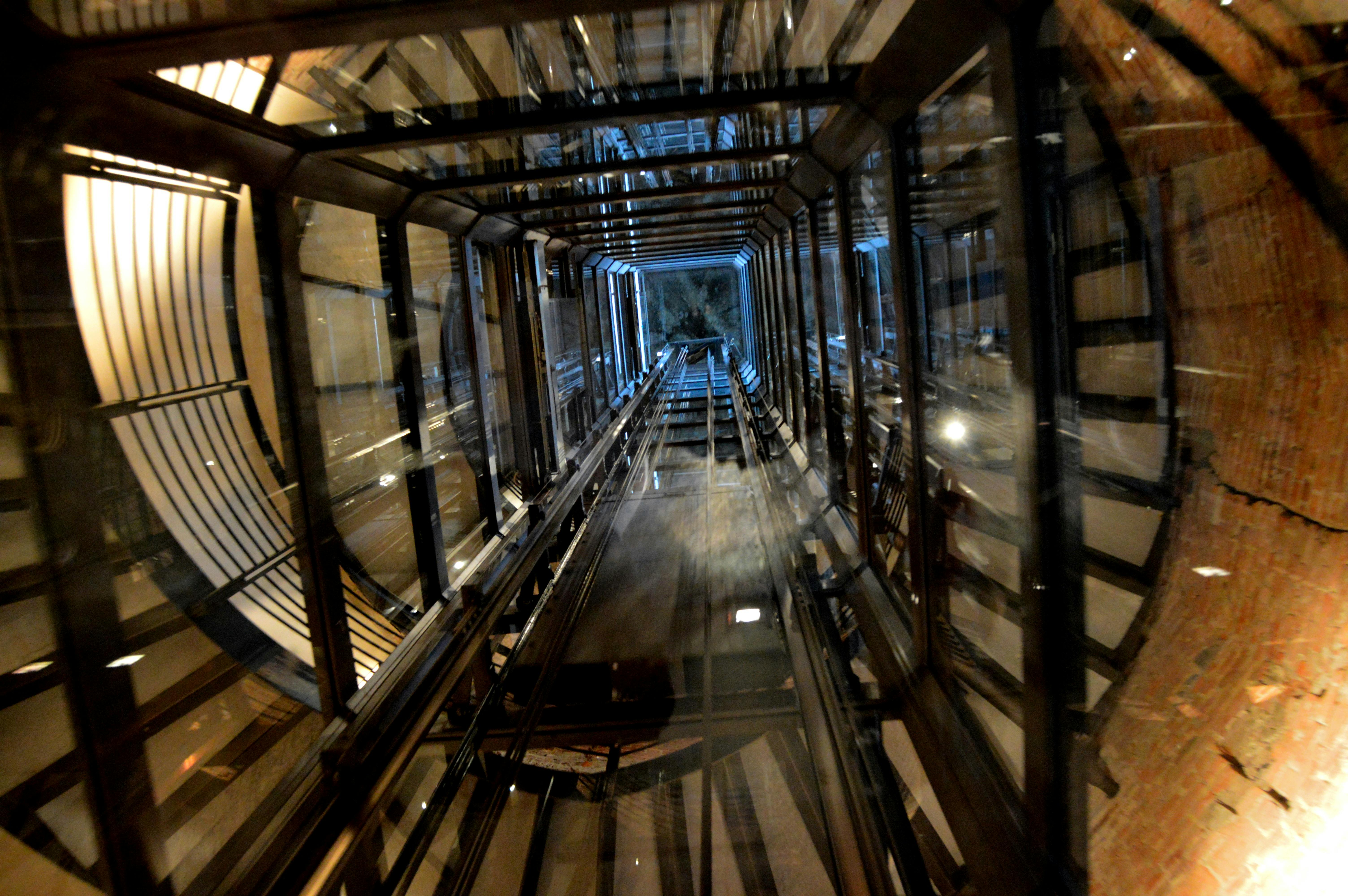 Blick nach oben durch einen detaillierten Aufzugsschacht aus Metall und Glas mit verschiedenen Reflexionen und Beleuchtungen.