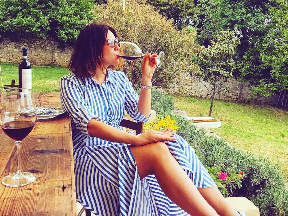 Woman in a striped dress sips wine at a wooden table outdoors, with a wine bottle and glass nearby, surrounded by greenery.