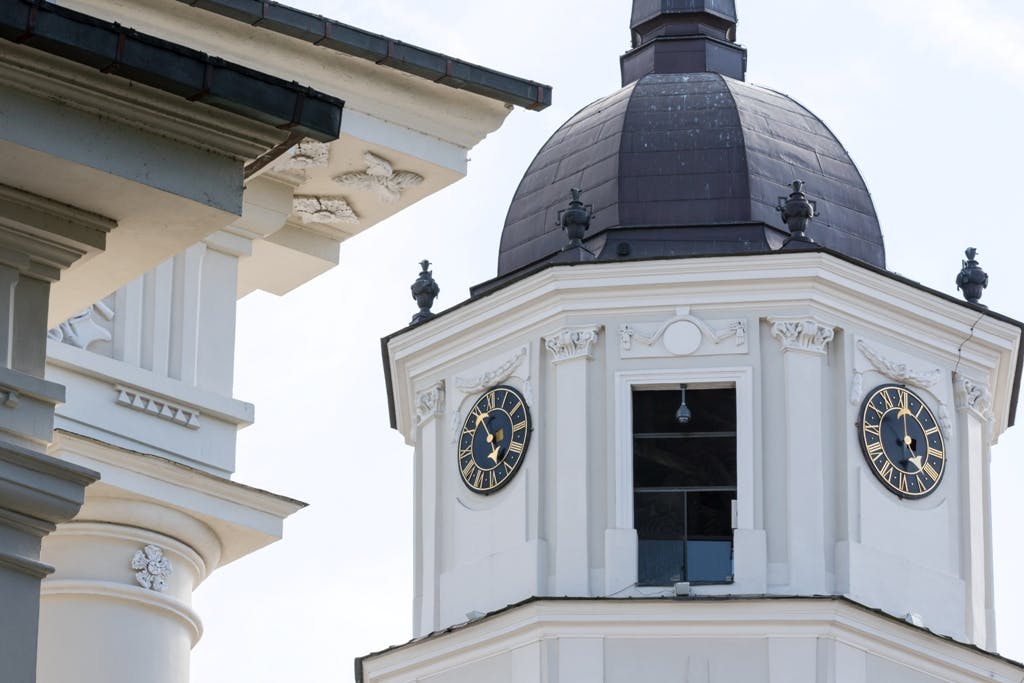 Close-up of a white clock tower with a dark dome roof, ornate detailing, and nearby architectural elements. The clock shows 4:50.