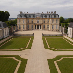 Ett storslaget slott med symmetriska trädgårdar, trimmade häckar, grusvägar och välskötta gräsmattor under en delvis molnig himmel.