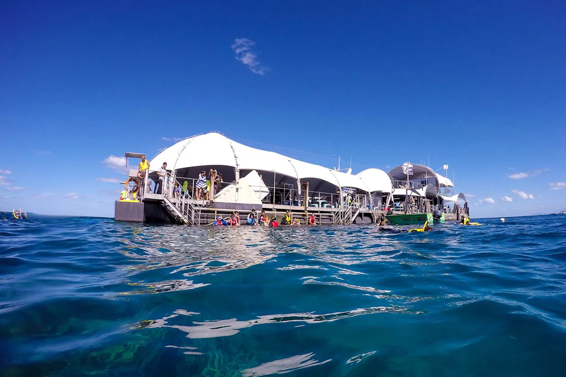 A floating platform with white tents and people preparing for water activities in a clear blue ocean under a sunny sky.