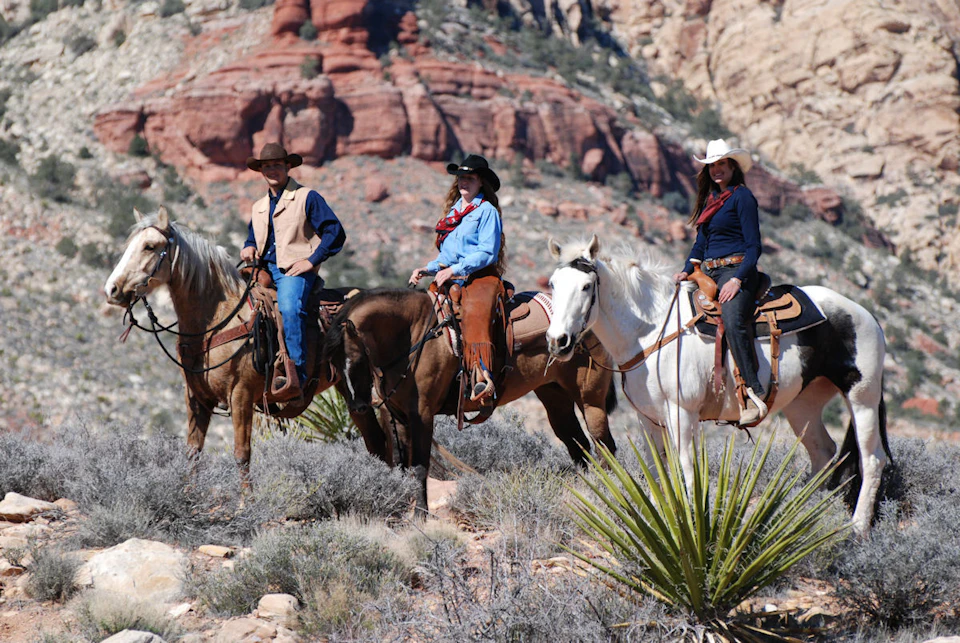 Three people in cowboy attire riding horses in a desert landscape with red rock formations in the background.